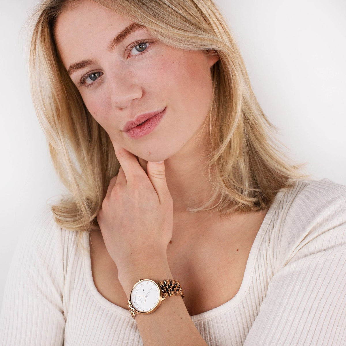 A blonde woman in a cream ribbed top touches her chin, the watch on her wrist.