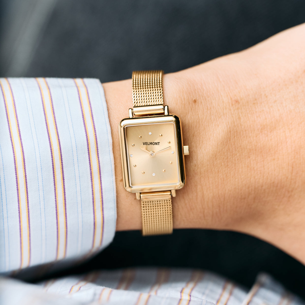 Gold-tone rectangular watch worn on a wrist with a striped shirt cuff.