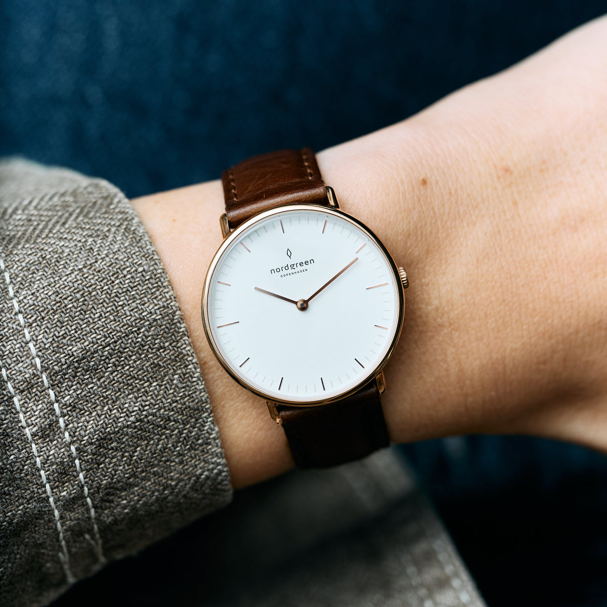 Rose gold-tone watch on a wrist beside a herringbone sleeve, dark brown strap.