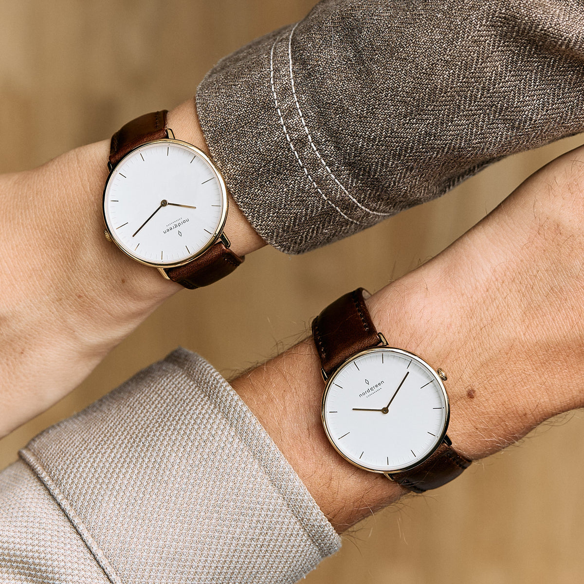 Two gold-tone watches with white dials and brown straps on crossed wrists.
