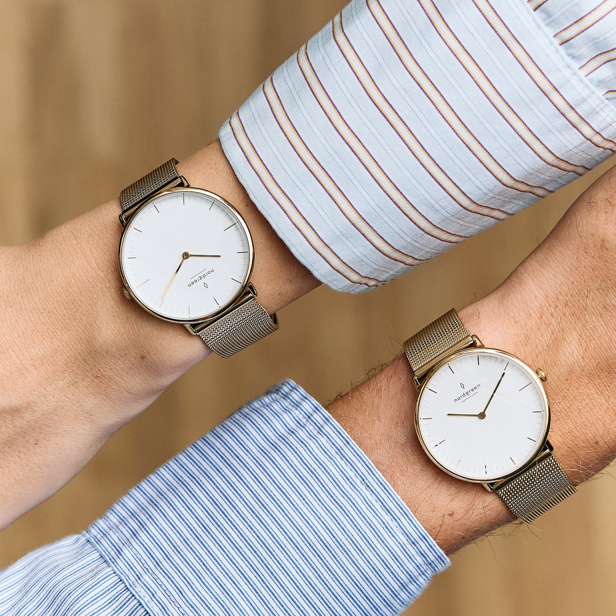 Two gold-tone mesh watches with white dials worn on crossed wrists in striped shirts.