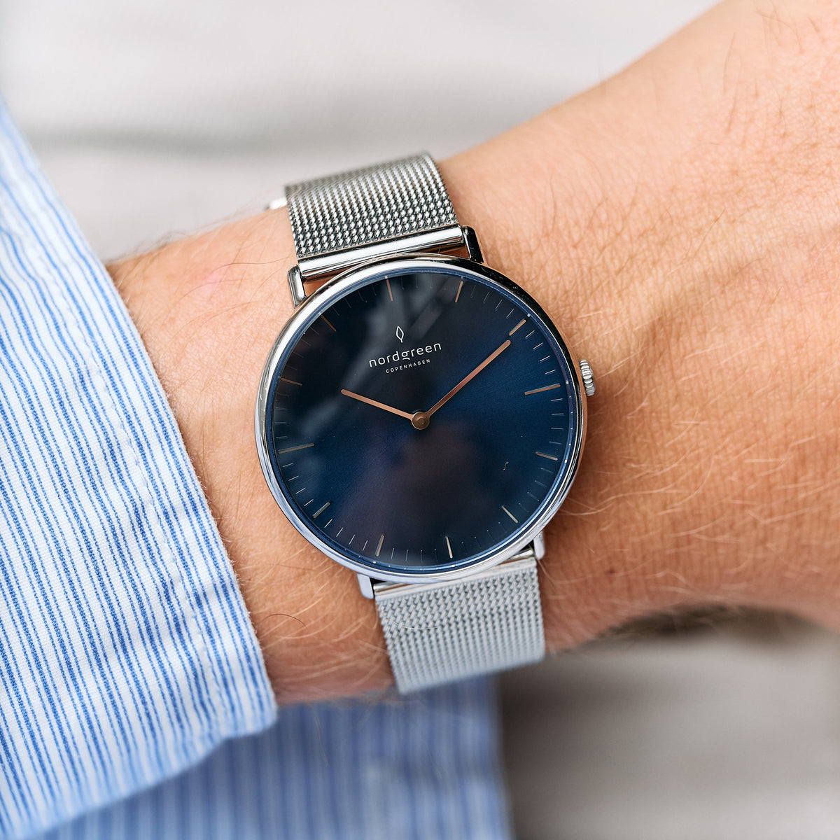 Navy dial with a silver-tone case and mesh strap, worn below a blue striped shirt cuff.