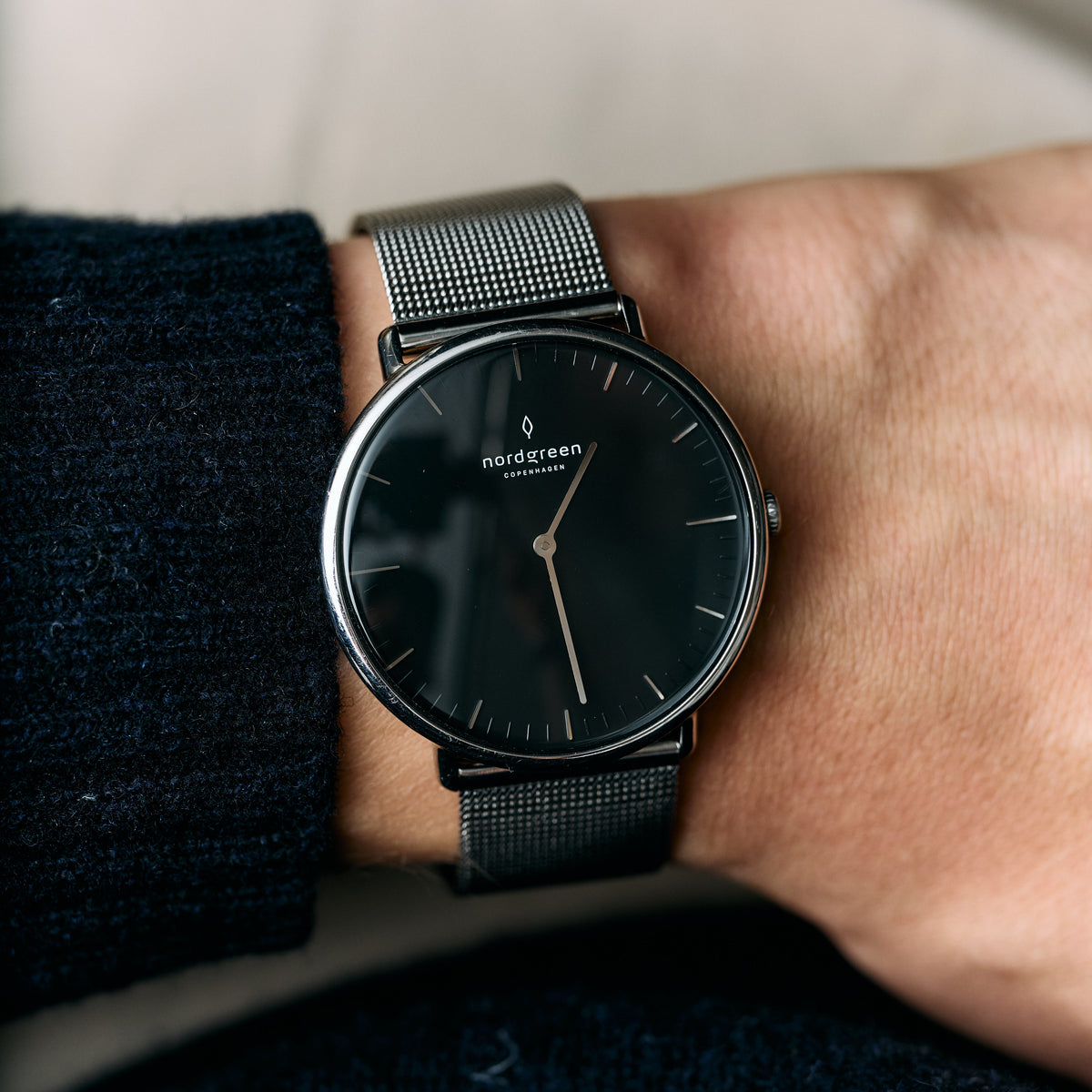 A black-dial watch on a dark mesh strap, worn beside a navy knit sleeve.