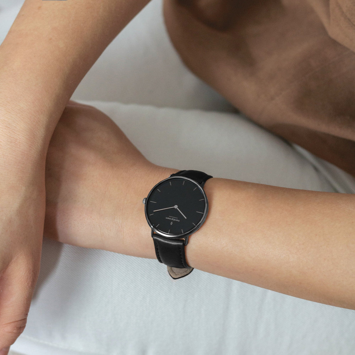 Arm resting on white bedding, wearing a dark-cased watch with a black dial and strap.