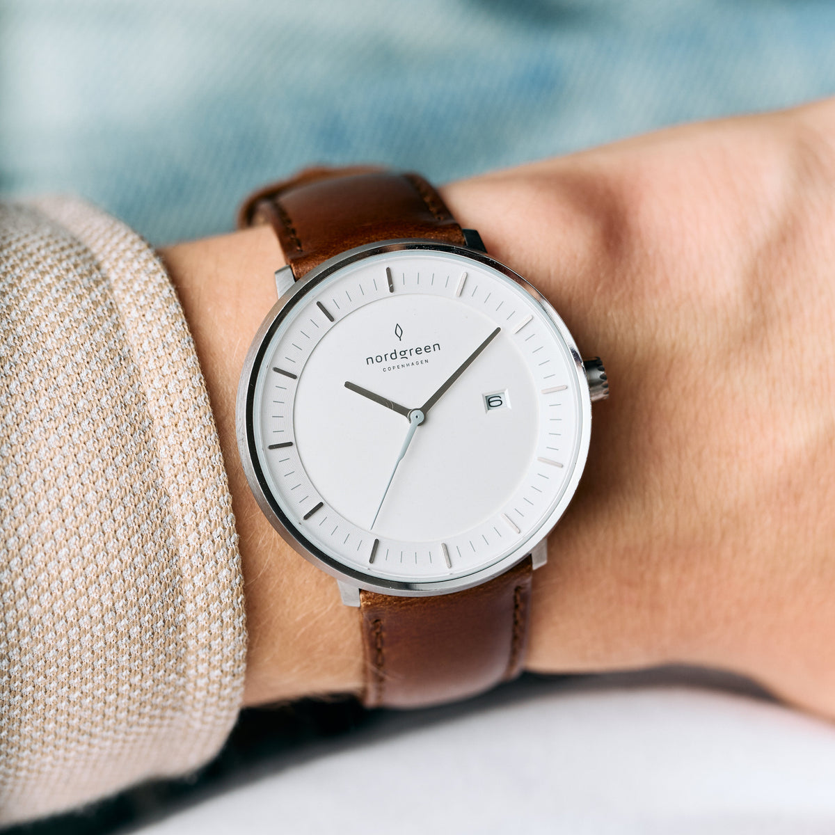 Brown strap and silver-tone case on a wrist with a beige knit sleeve, white dial facing up.