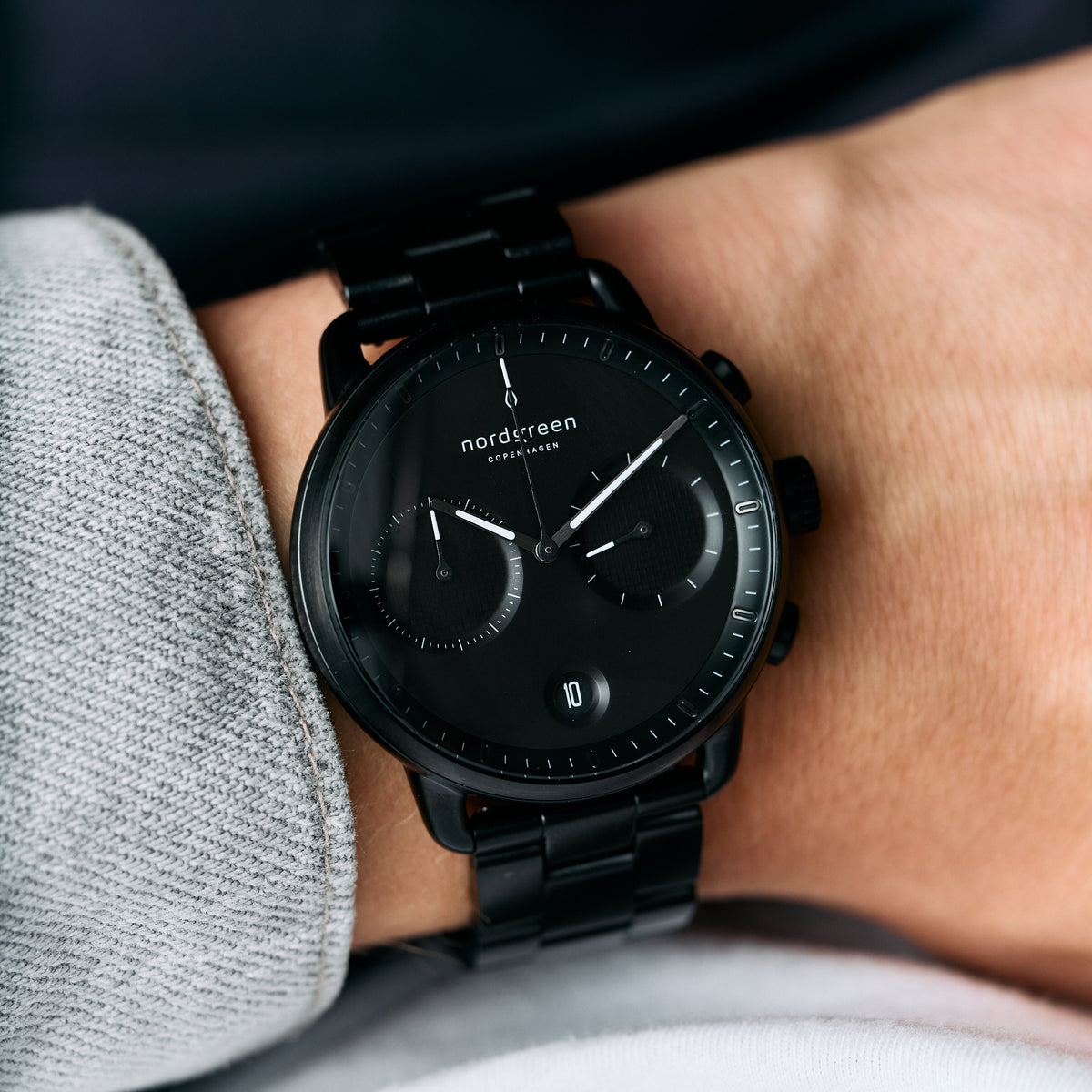 The black bracelet watch worn on a wrist beside a gray sleeve.
