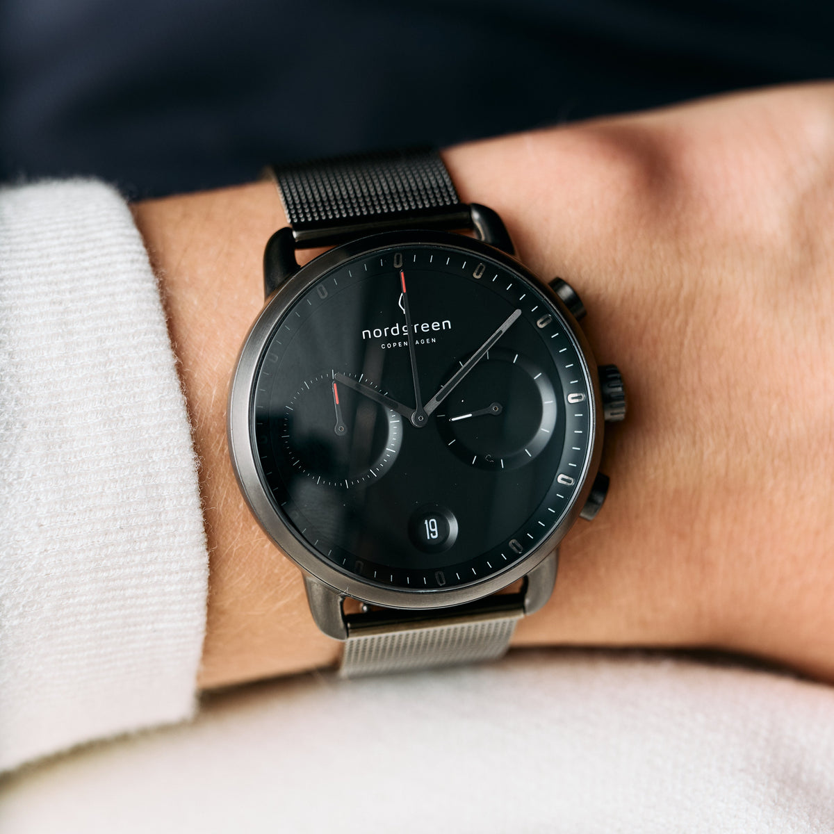 The dark gunmetal watch worn on a wrist beside a white knit sleeve.