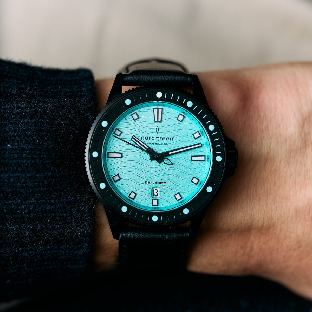 The turquoise-dial watch on a wrist beside a dark knit sleeve.