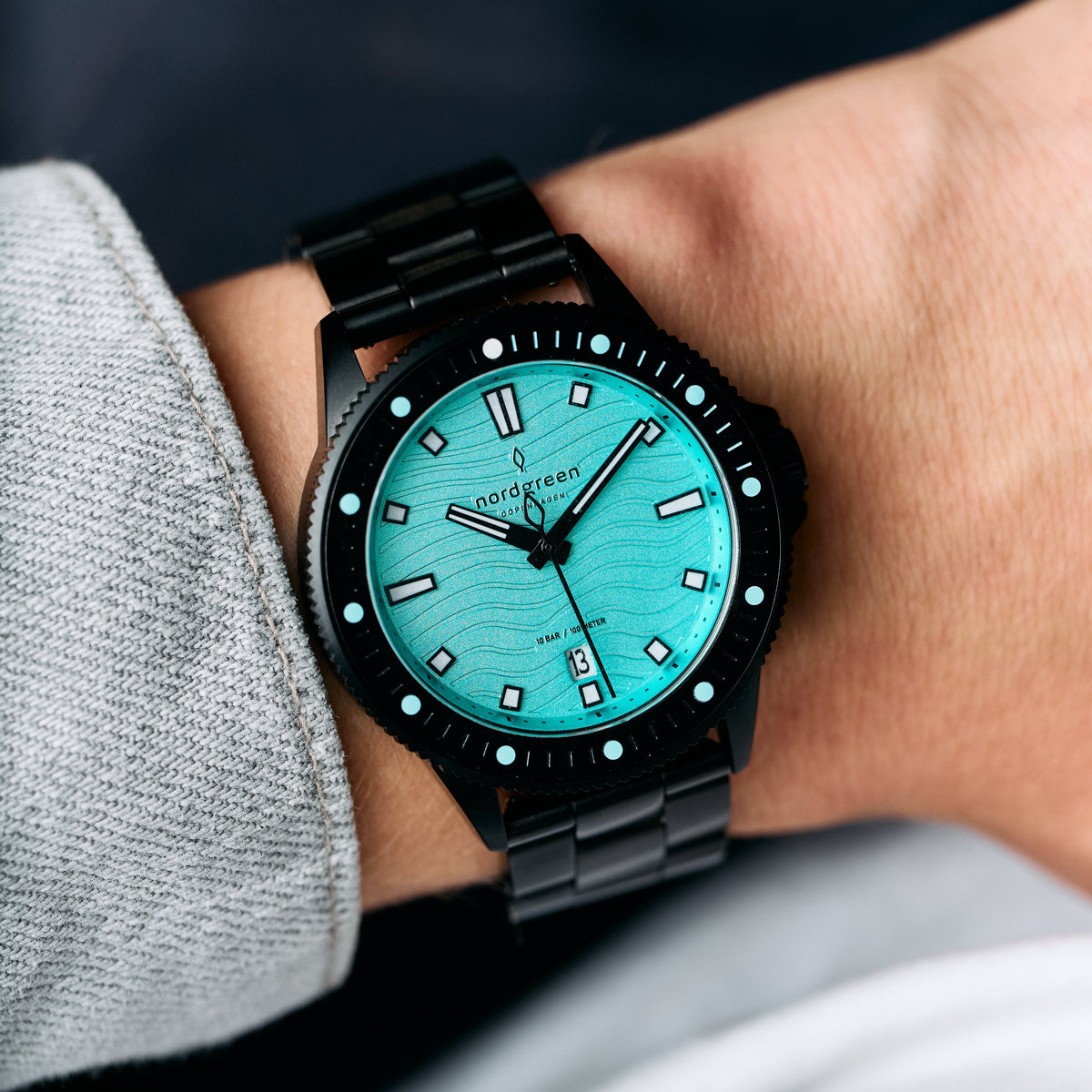 The turquoise dial watch worn on a wrist beside a gray sleeve.