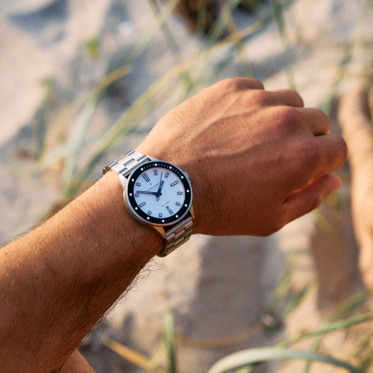 The white-dial watch on a silver-tone link bracelet, outdoors among beach grass.