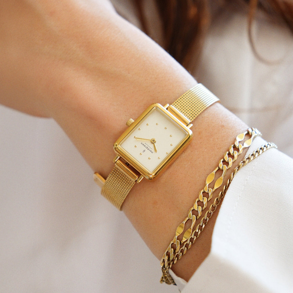 Gold-tone watch stacked with chain bracelets on a wrist in a white sleeve.