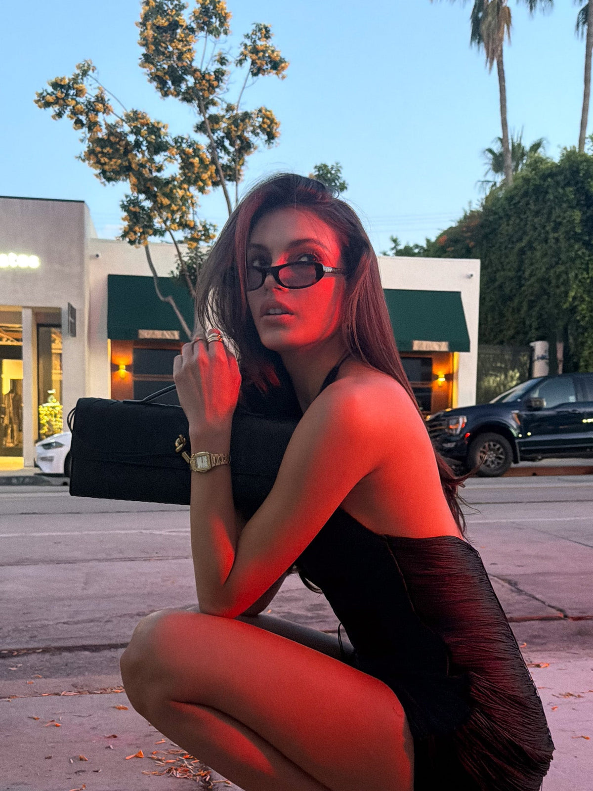 Woman crouching on a city street at dusk wearing the sunglasses