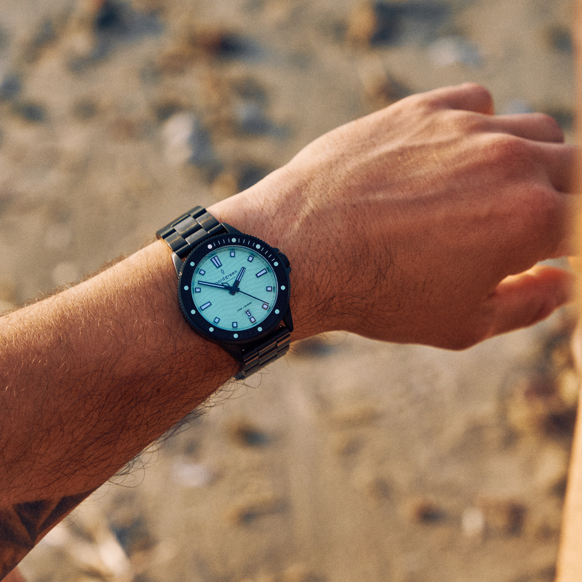 Worn outdoors on a wrist above a sandy beach, black bracelet visible.