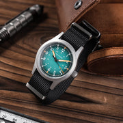 Teal blue dial field watch on a wooden desk next to a pen