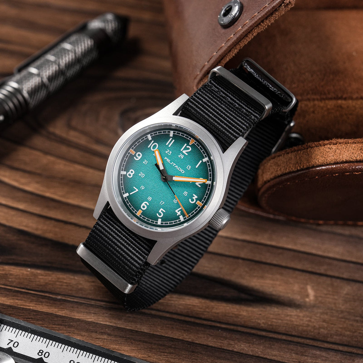 Teal blue dial field watch on a wooden desk next to a pen