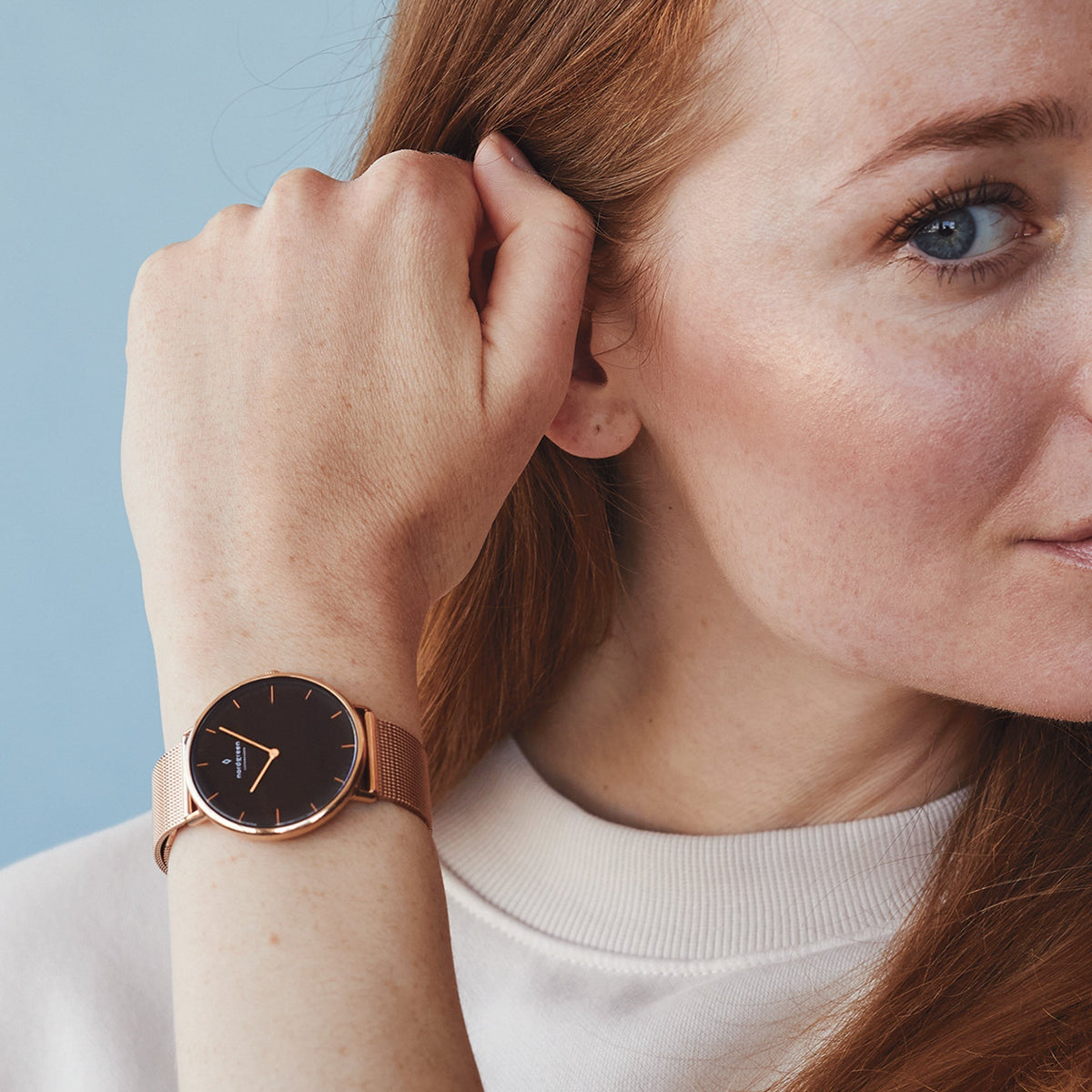 A red-haired woman in a cream sweater raises her hand, showing the black-dial watch.