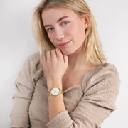 A woman in a beige textured top rests her chin on her hand, wearing the gold-tone watch.