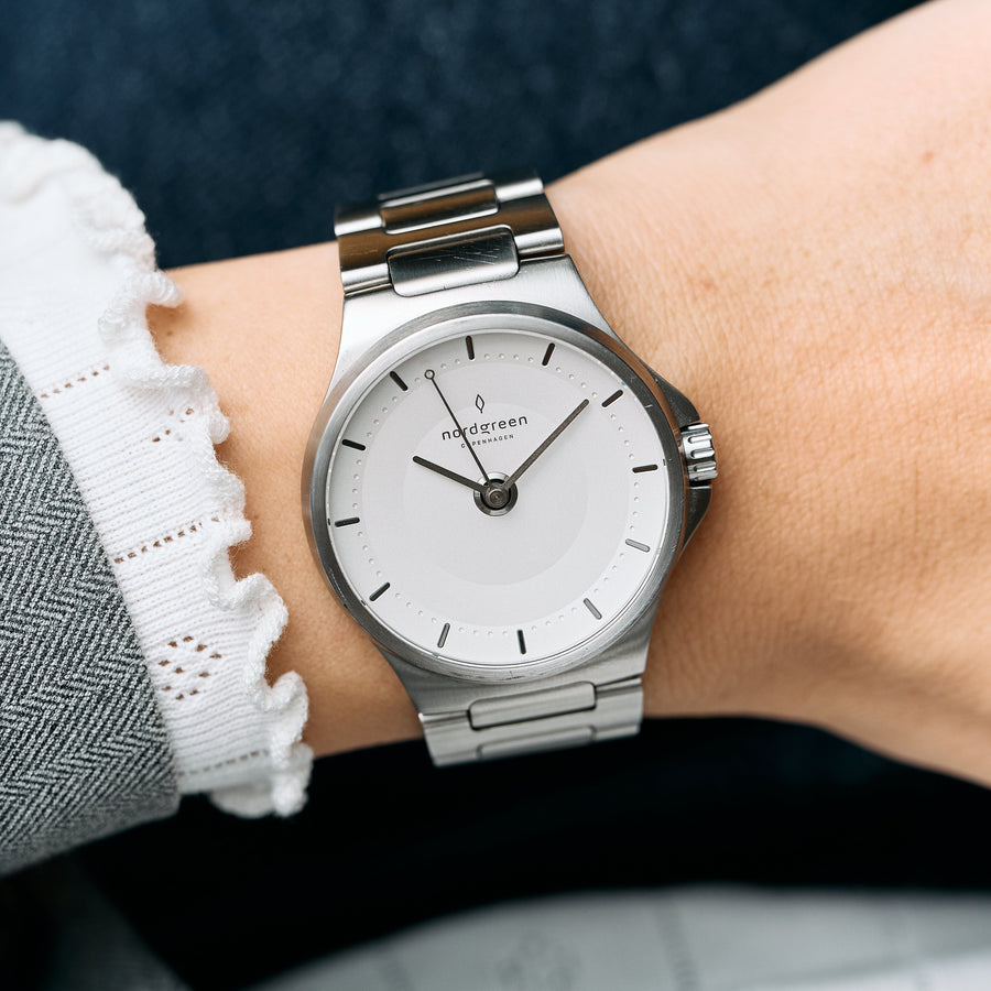 The silver-tone watch worn on a wrist with a white lace cuff and gray coat.