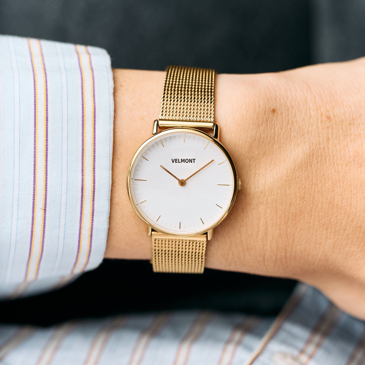 Gold-tone mesh watch with a white dial, worn below a striped shirt cuff.