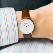 A white-dial watch with a silver-tone case worn under a striped shirt cuff on a brown strap.
