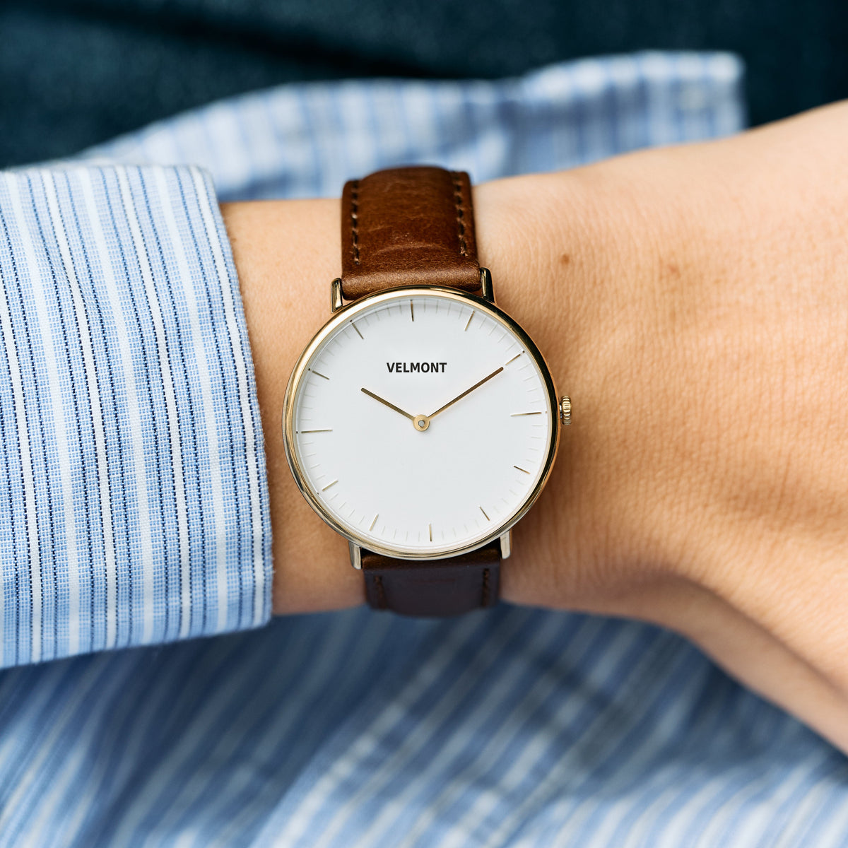 The gold-tone watch worn on a wrist with a blue striped shirt cuff.