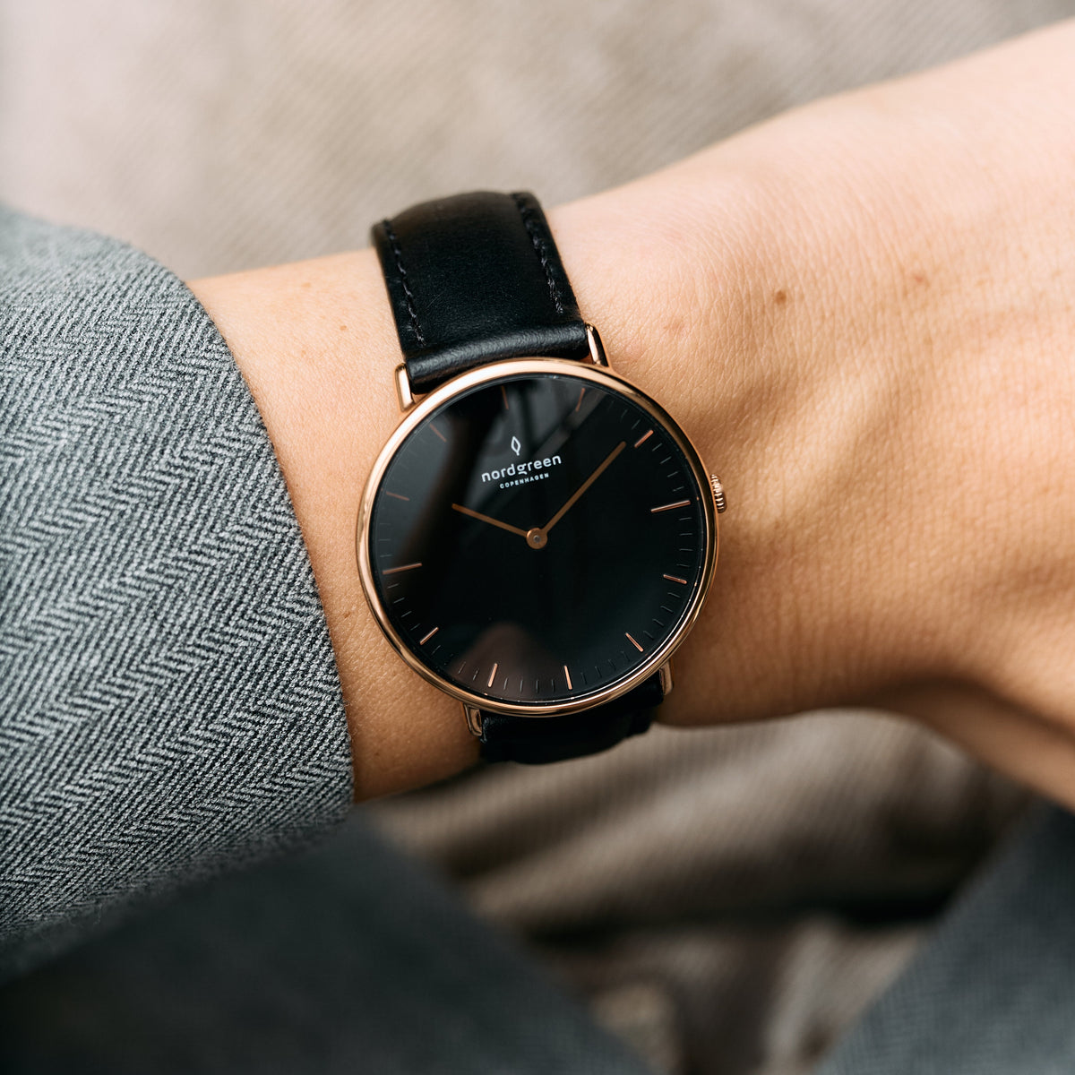 Rose gold-tone case with a black dial on a black strap, shown on a wrist by a herringbone sleeve.