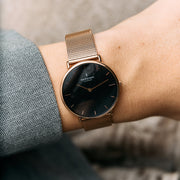 Black dial with a rose gold-tone case and mesh, worn against a gray herringbone sleeve.