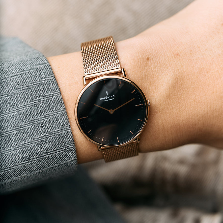 Black dial with a rose gold-tone case and mesh, worn against a gray herringbone sleeve.