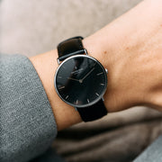 A silver-tone watch with a black dial and black strap, worn with a gray herringbone sleeve.