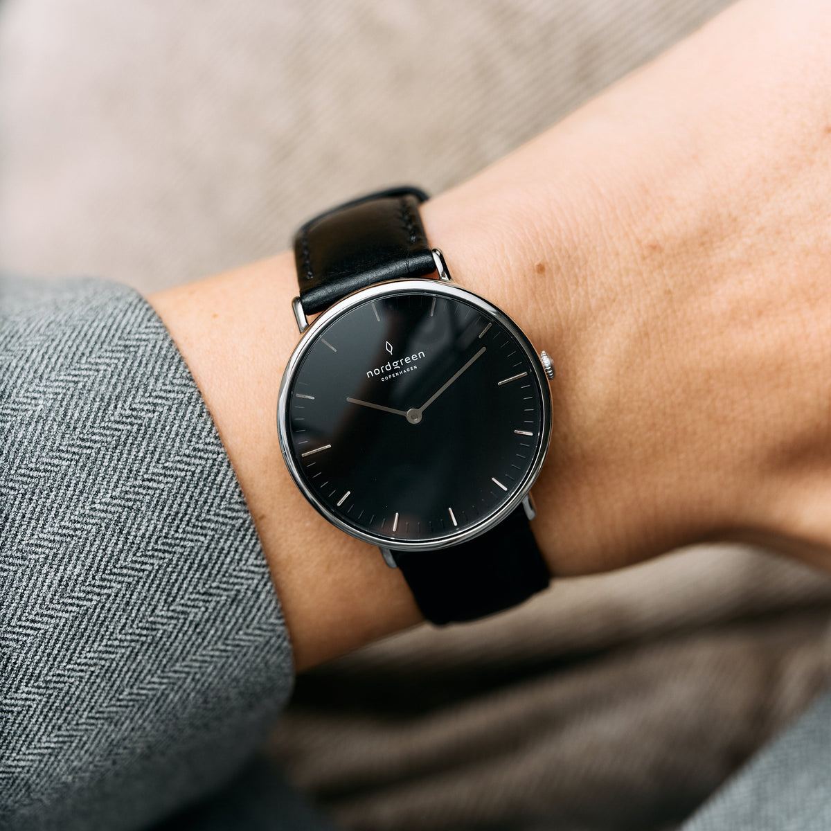 A silver-tone watch with a black dial and black strap, worn with a gray herringbone sleeve.