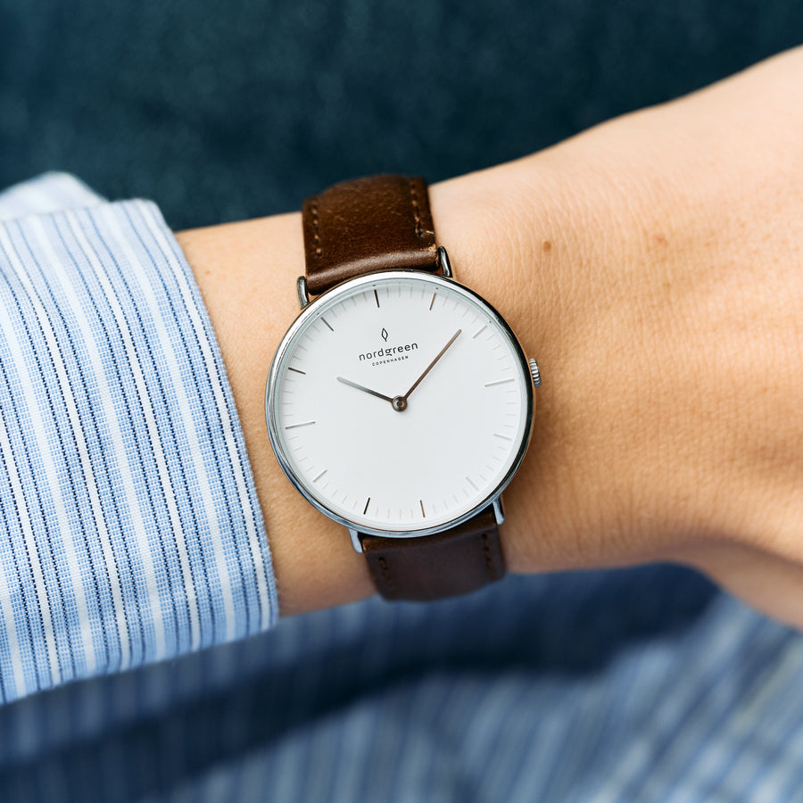 The watch on a wrist with a blue striped shirt, showing a dark brown strap.