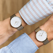 Two gold-tone mesh watches with white dials worn on crossed wrists in striped shirts.
