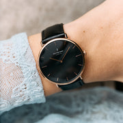 The watch worn on a wrist beside a pale blue lace cuff, black dial and rose gold-tone case.