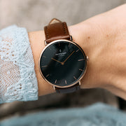 Black dial and rose gold-tone case on a brown strap, worn next to a pale blue lace cuff.