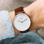 The watch on a wrist with a pale blue lace sleeve and brown strap.