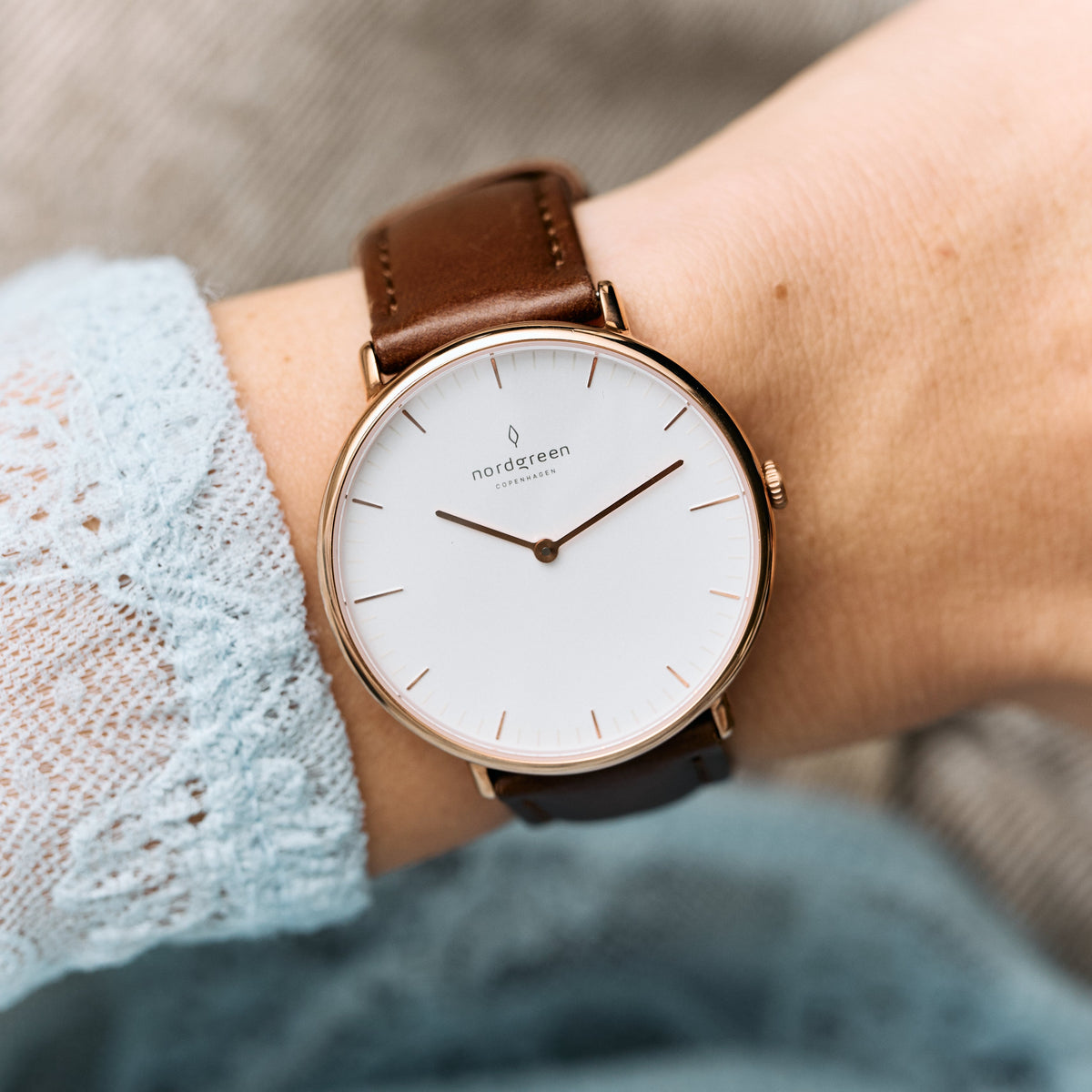 The watch on a wrist with a pale blue lace sleeve and brown strap.