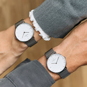 Two white-dial watches on dark mesh straps worn on crossed wrists.