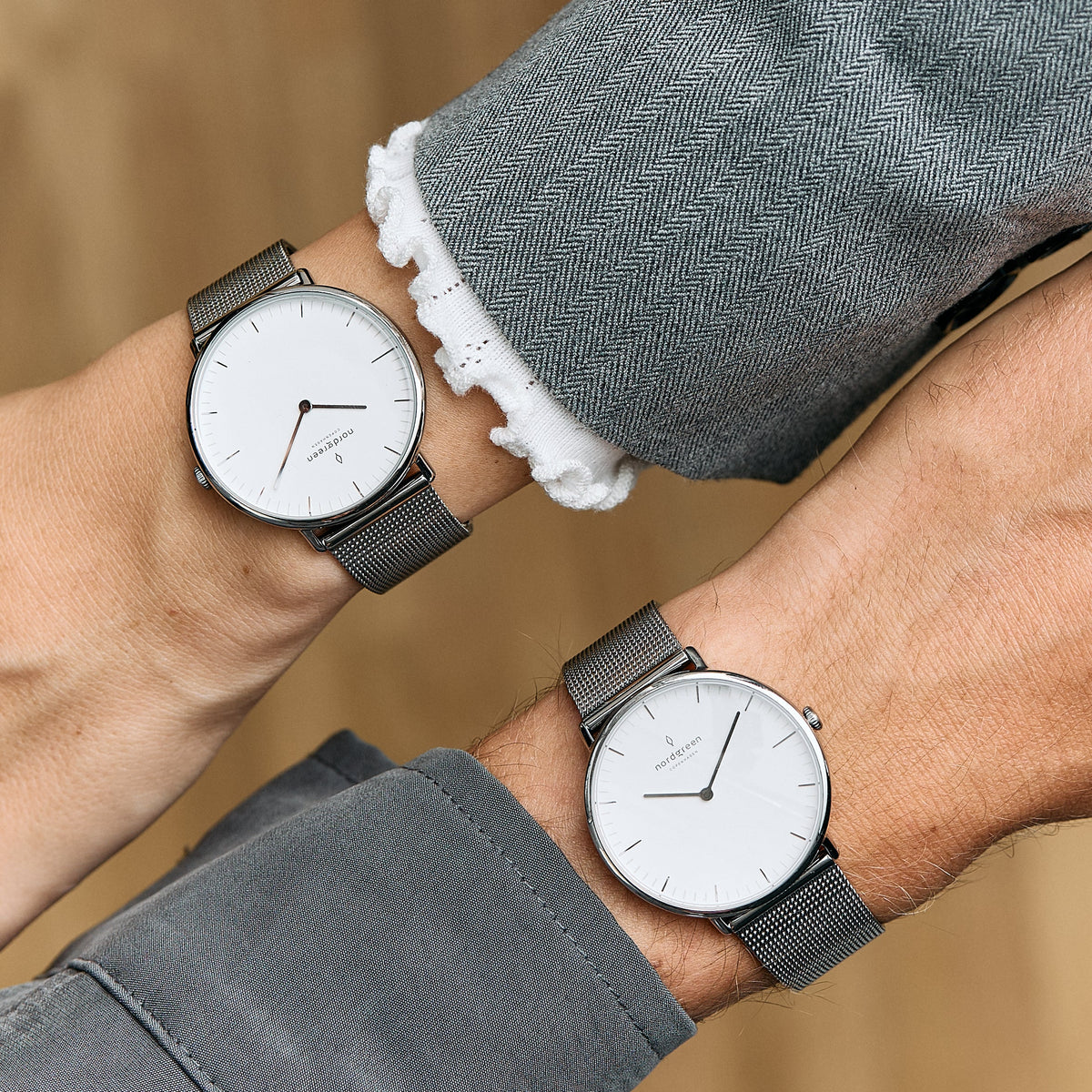 Two white-dial watches on dark mesh straps worn on crossed wrists.