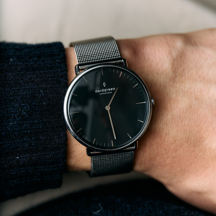 A black-dial watch on a dark mesh strap, worn beside a navy knit sleeve.