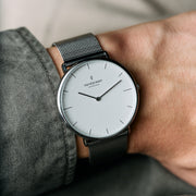White-dial watch on a dark mesh strap, worn with a gray shirt cuff.
