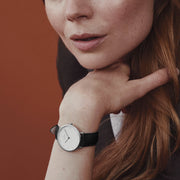A woman resting her chin on her hand, wearing the white-dial watch on a black strap.