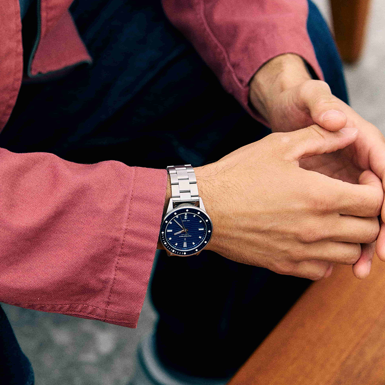 Hands clasped on a table, the blue dial watch on a man in a red shirt.
