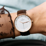 Rose gold-tone case and white dial on a dark brown strap, beside a sheer dotted sleeve.