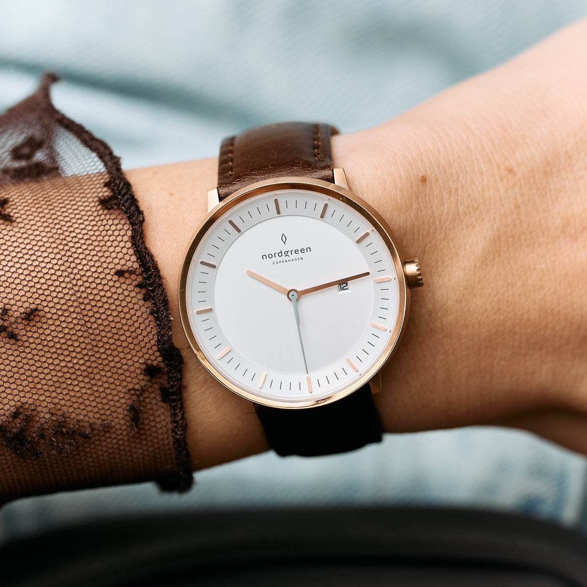 Rose gold-tone case and white dial on a dark brown strap, beside a sheer dotted sleeve.
