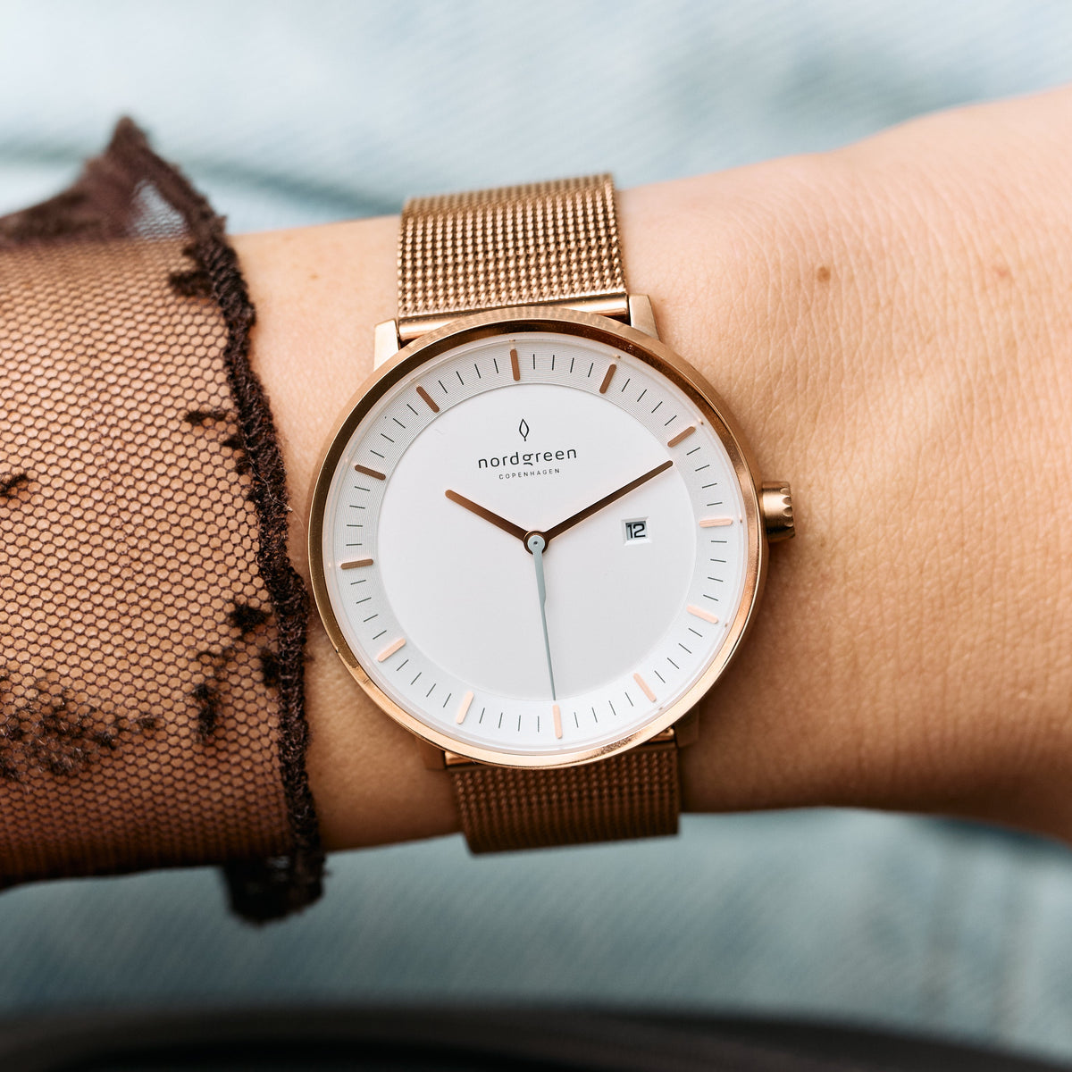 On the wrist beside a sheer dotted sleeve, a white dial in a rose gold-tone case and mesh.