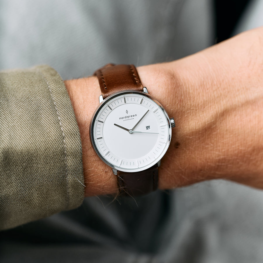 A white-dial watch on a brown strap, worn with an olive sleeve rolled back.