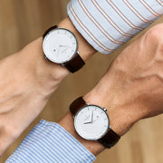 Two crossed wrists in striped shirts, each with a silver-tone watch on a dark brown strap.