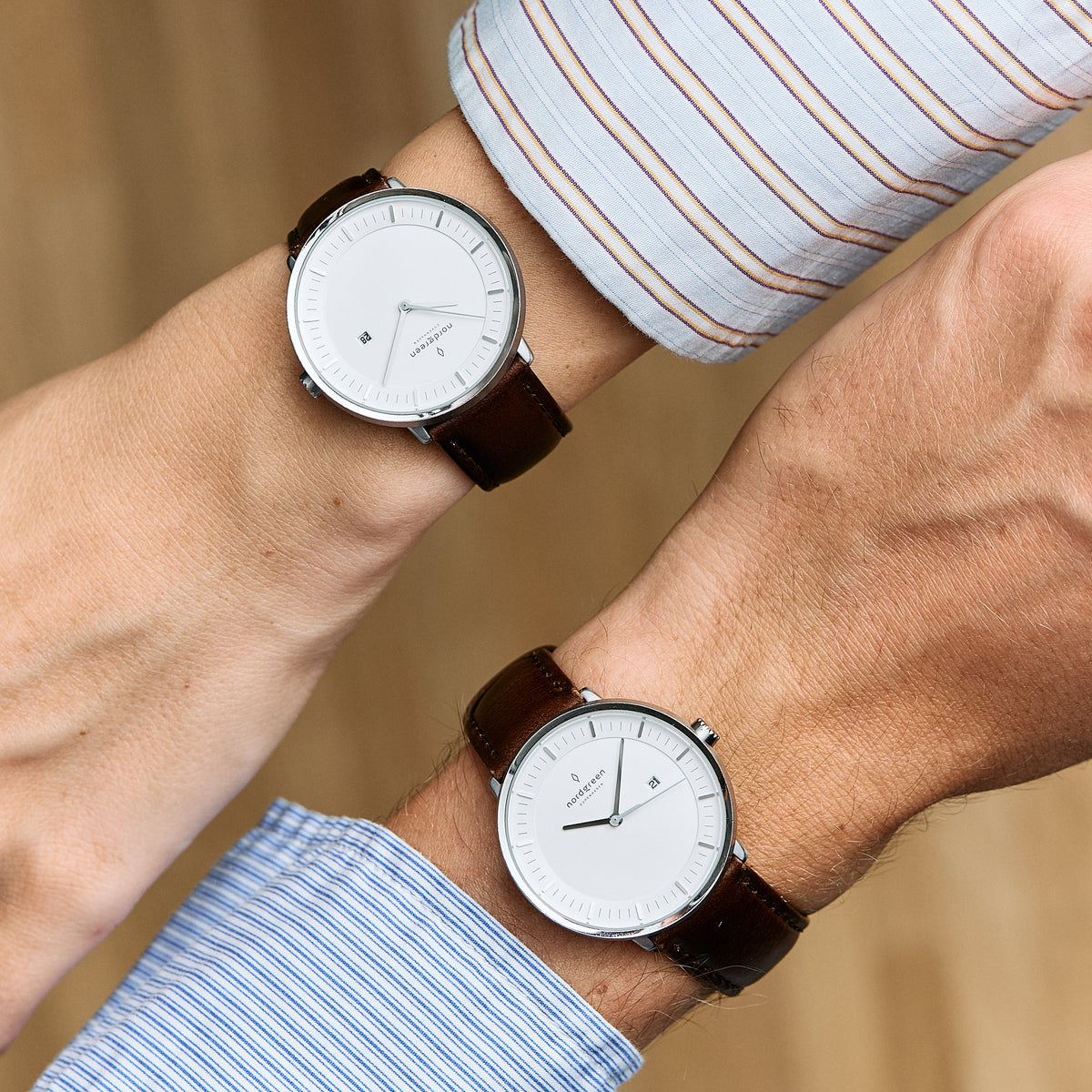 Two crossed wrists in striped shirts, each with a silver-tone watch on a dark brown strap.