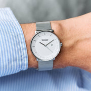 Silver-tone mesh watch on a wrist with a blue pinstripe shirt cuff, white dial visible.
