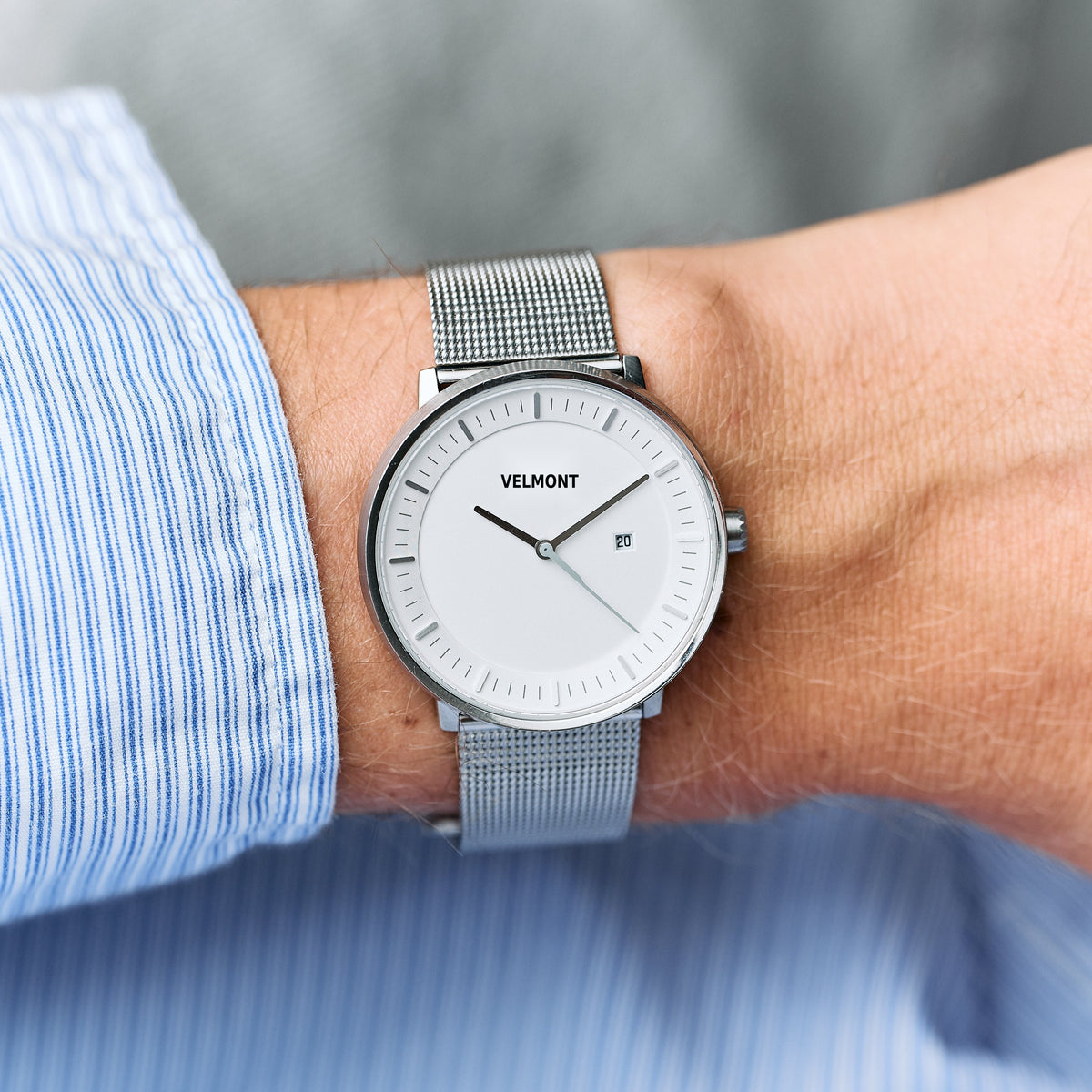 Silver-tone mesh watch on a wrist with a blue pinstripe shirt cuff, white dial visible.